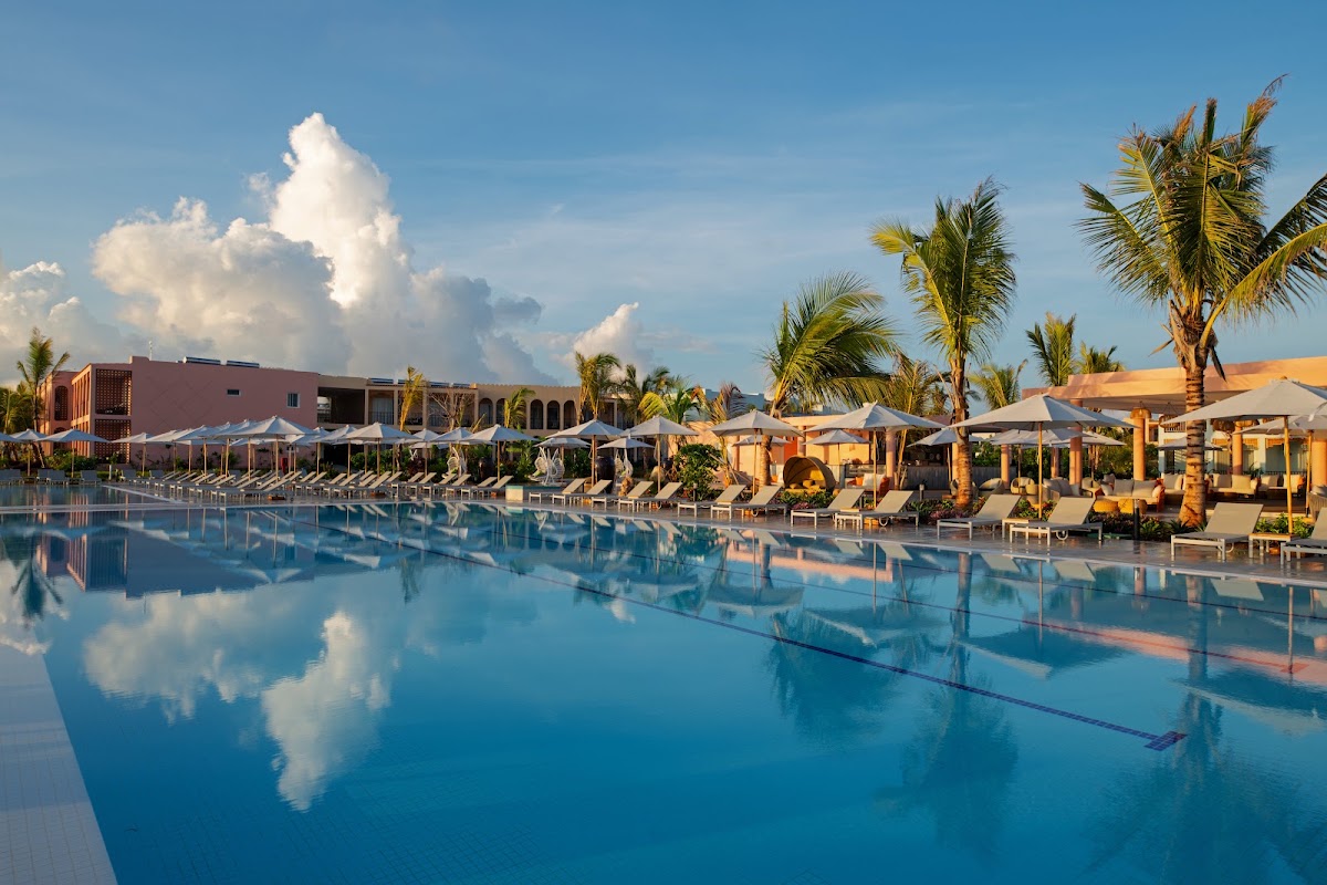 Large resort pool with white umbrellas, lounge chairs, and palm trees lining the deck, with a terracotta building and cabanas under a blue sky.
