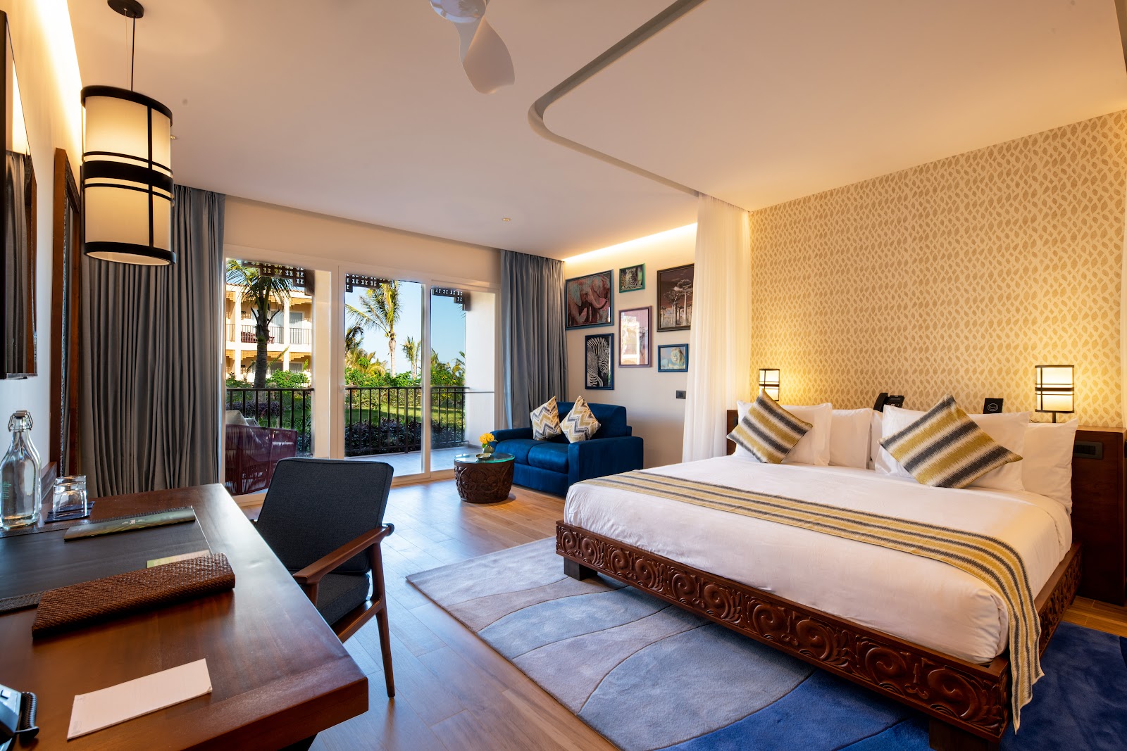 Spacious resort room with king bed, blue seating area, and balcony overlooking palm trees and ocean at The Mora Zanzibar.