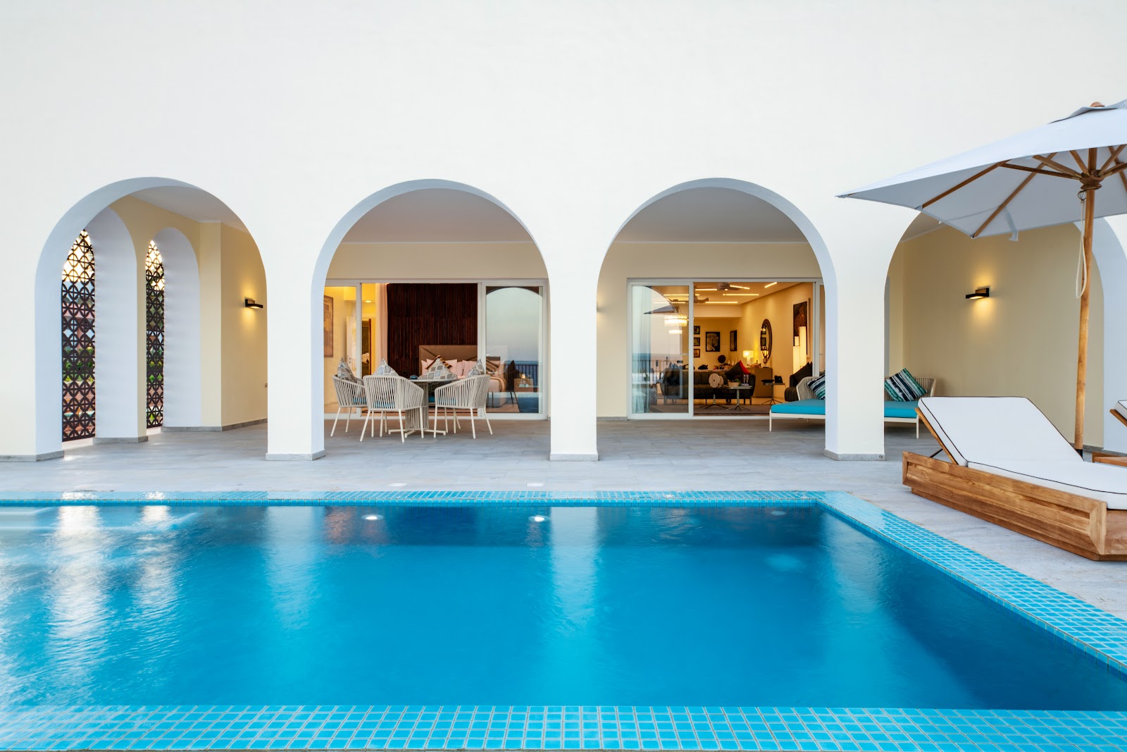 A blue swimming pool reflects white arched colonnades at The Mora Zanzibar, with lounge chairs and dining areas visible through open archways.