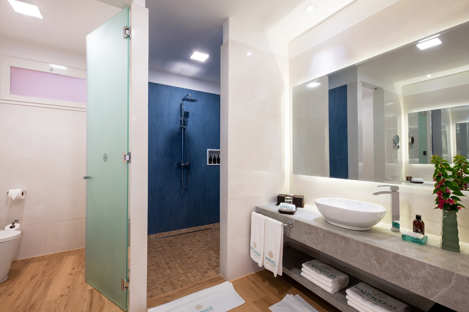 Modern bathroom with glass-enclosed shower with blue tile walls, white vessel sink, illuminated mirror, and wood flooring.