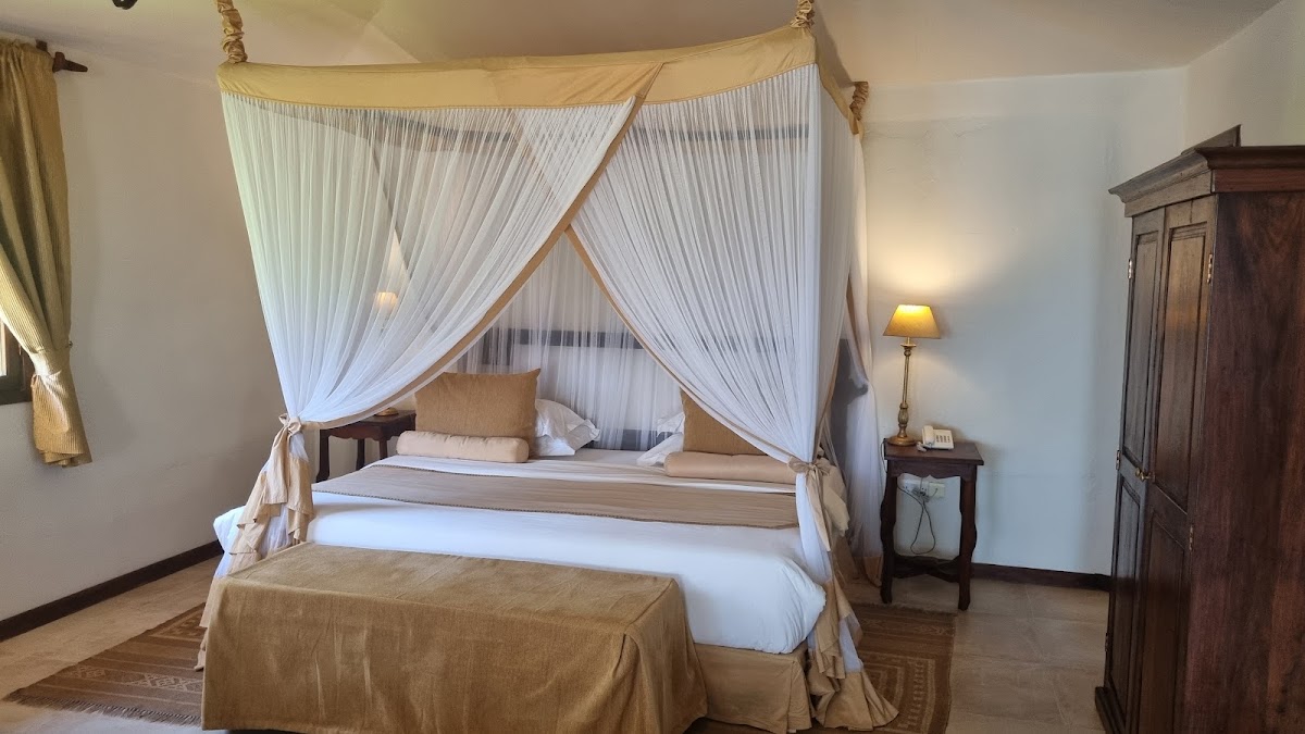 Four-poster canopy bed with white sheer curtains and gold accents in a spacious resort bedroom with dark wood furnishings.