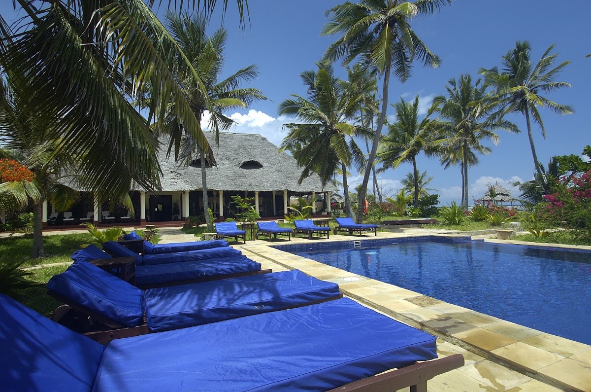Blue loungers line the pool deck at The Palms Zanzibar, with a thatched-roof main building and palm trees under clear sky.