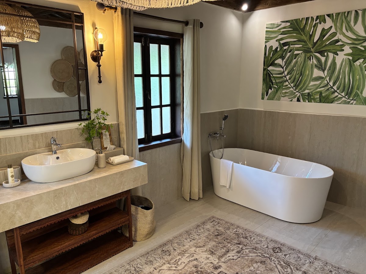 Spacious bathroom at The Palms Zanzibar with white oval bathtub, vessel sink, tropical leaf artwork, and natural light from glass doors.
