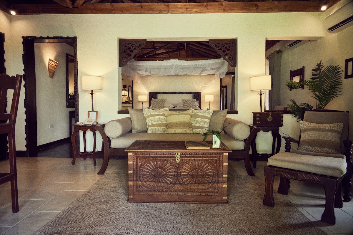 Spacious resort suite with canopy bed, cream sofa, carved wooden furniture, and tropical plants beneath exposed beam ceiling.