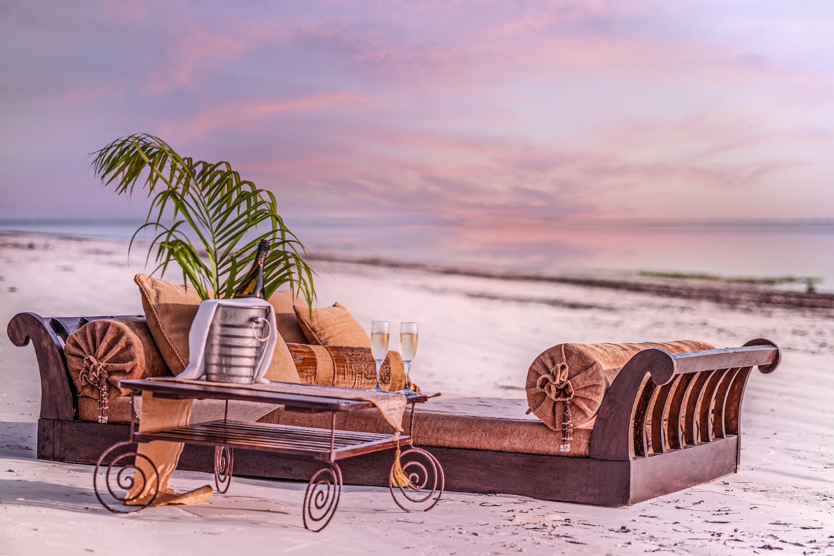 Ornate lounge seating with cushions and a potted palm plant on a beach at sunset, with champagne glasses and gentle waves in the background.