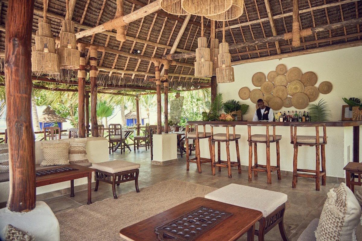 Open-air bar and lounge with thatched roof, wooden columns, woven pendant lights, and a bartender behind the counter at The Palms Zanzibar.