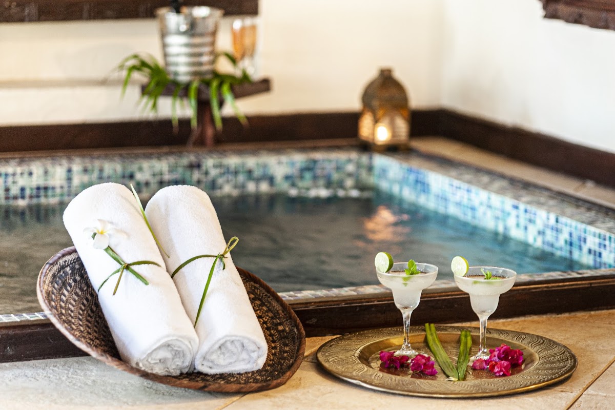 Rolled white towels with grass ties in a woven basket beside a small pool, with two margaritas and pink flowers on a brass plate nearby.