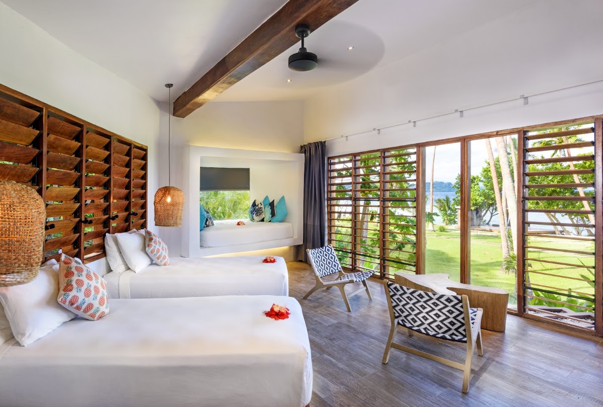 Modern resort bedroom with two queen beds, wooden beams, woven pendant light, and louvered doors opening to tropical grounds and ocean view.