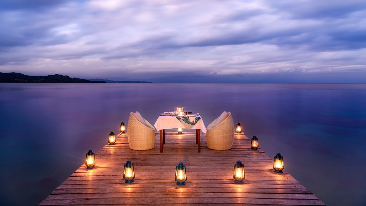 Wooden pier at dusk with two woven chairs, a dining table set for two, and lit lanterns surrounding the setup on calm ocean water under a purple twilight sky.