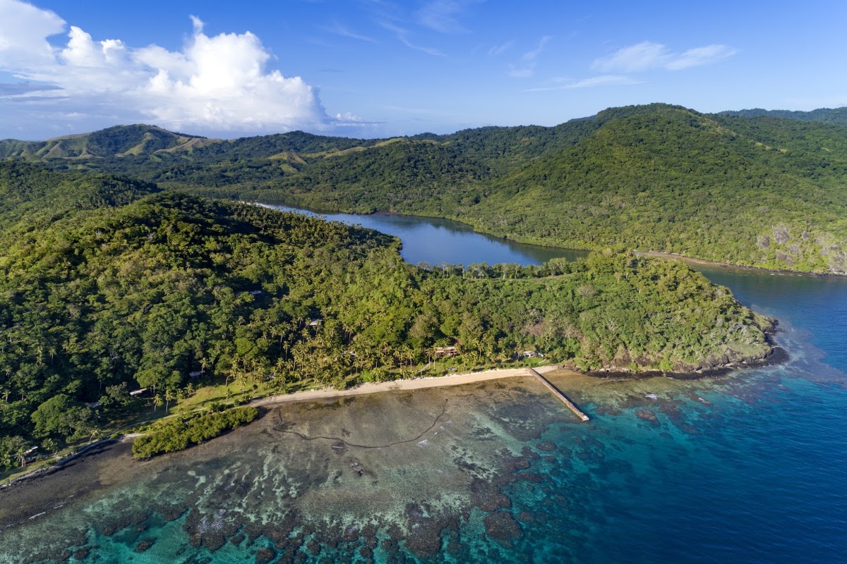 Aerial view of The Remote Resort Fiji Islands nestled between dense jungle, a sandy beach, coral reef, and inland lagoon under blue sky.