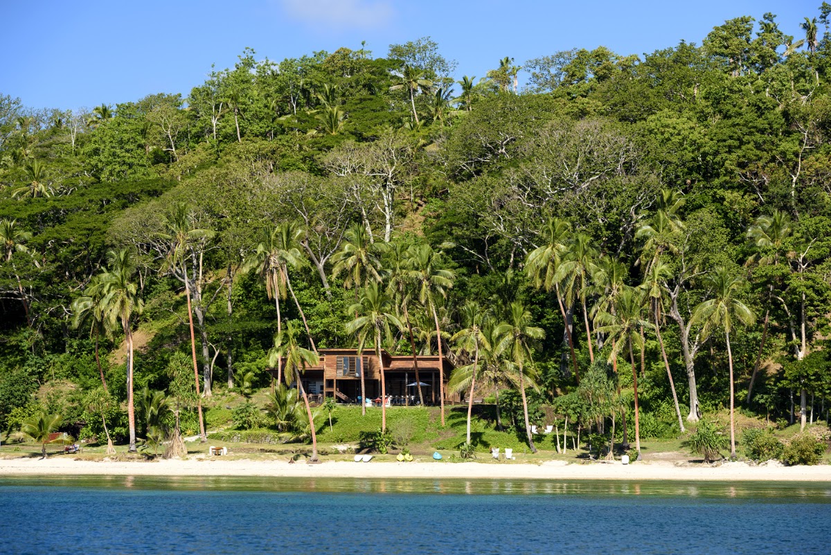 Wooden lodge nestled on a hillside surrounded by dense tropical forest and palm trees, with white sand beach and blue water in foreground.