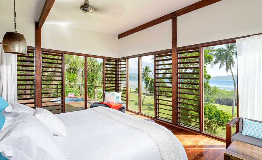 Bedroom with white bedding and wooden beams overlooks palm trees, manicured grounds, and ocean views through louvered glass doors at The Remote Resort Fiji Islands.