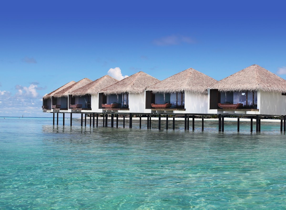 Row of overwater bungalows with thatched roofs at The Residence Maldives, set in turquoise lagoon water under clear blue sky.