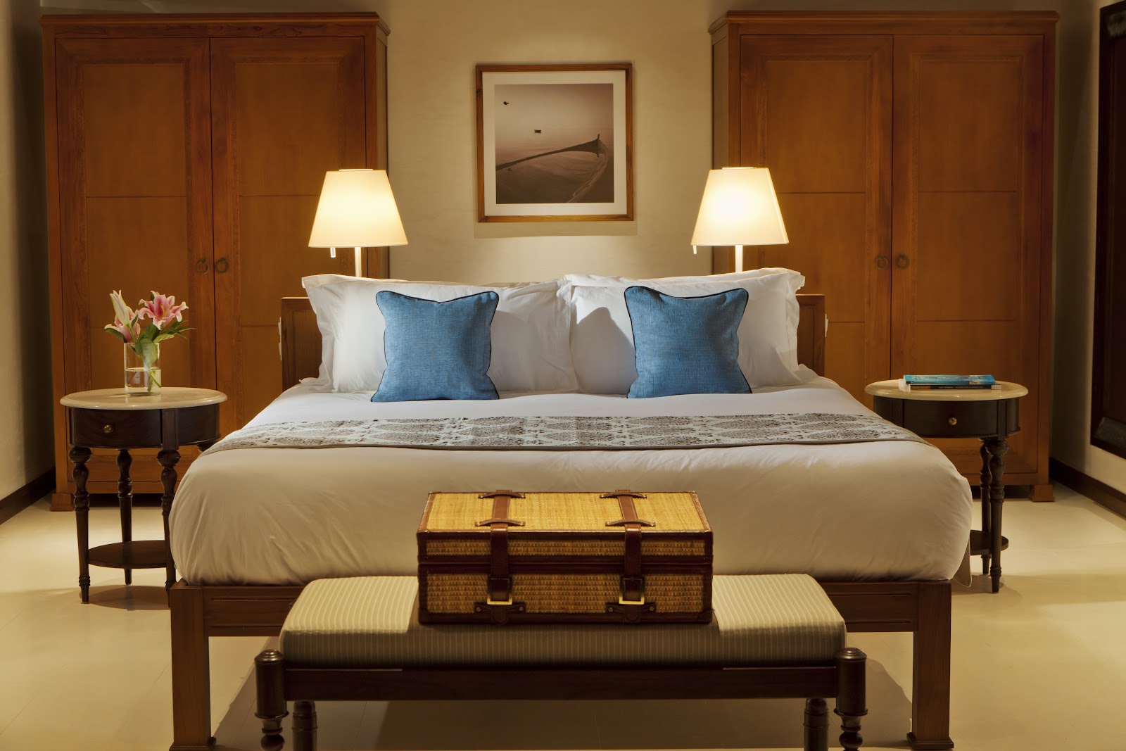 Resort bedroom with king bed, blue pillows, wooden wardrobes, side tables with lamps, and woven trunk at foot of bed.