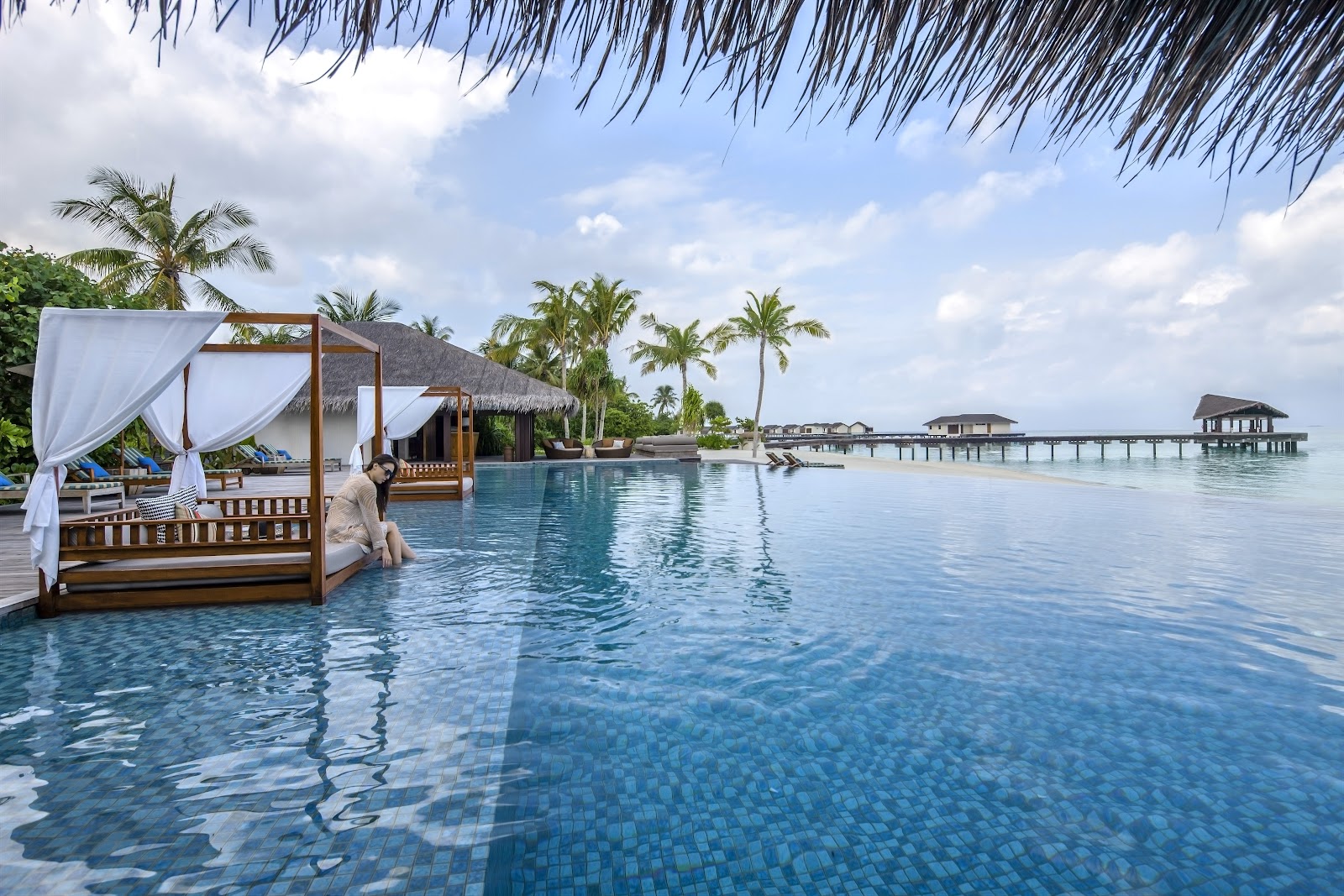 Wooden cabana bed with white curtains sits in an infinity pool overlooking overwater bungalows and palm trees at The Residence Maldives.