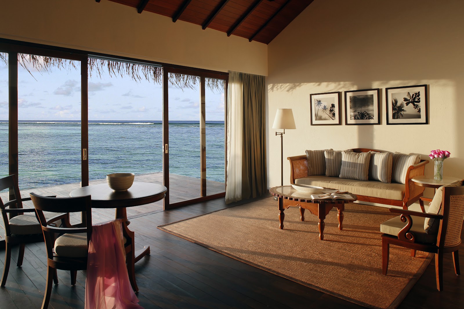 Overwater bungalow suite with floor-to-ceiling glass doors opening to the Indian Ocean, wood-beamed ceiling, striped sofa, and wooden furniture.