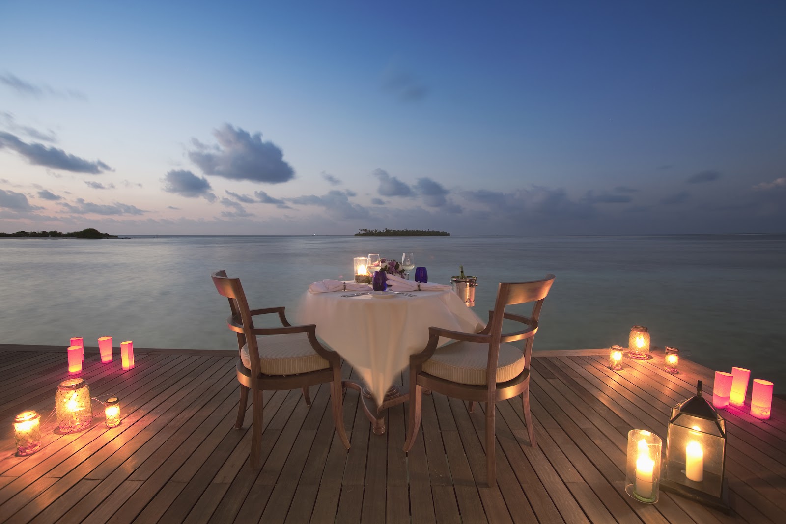 Candlelit dinner table set for two on a wooden deck overlooking calm lagoon waters at dusk with distant islands.