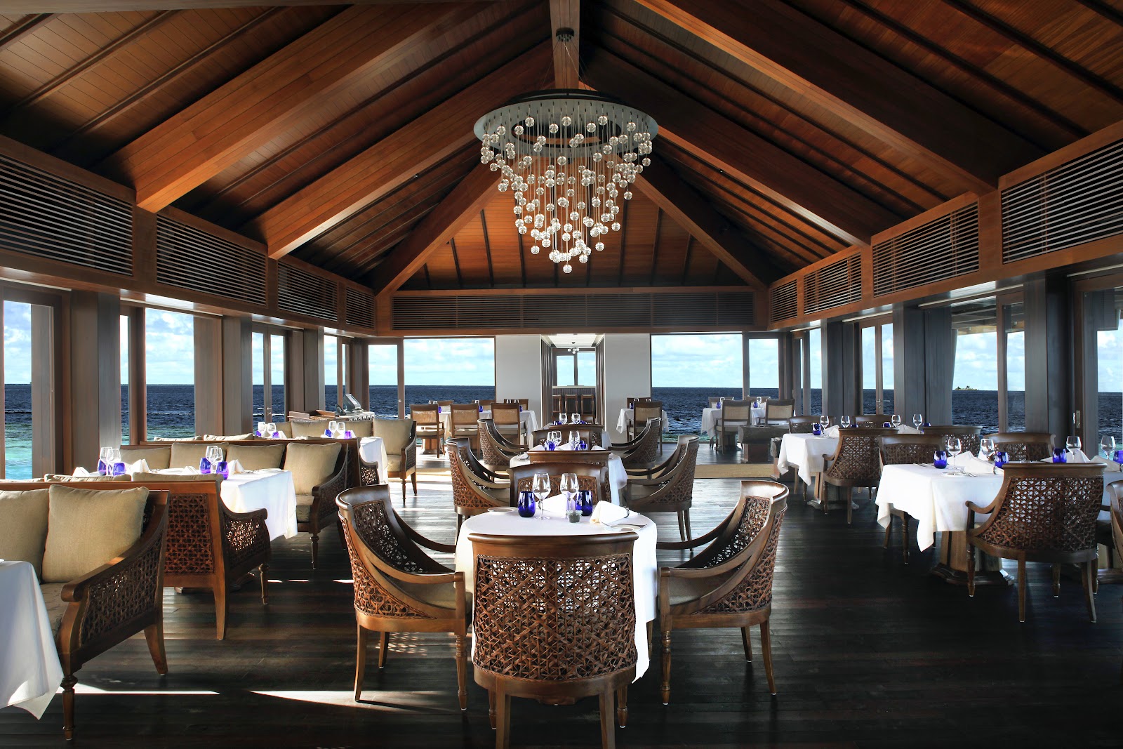 Fine dining restaurant with wooden ceiling beams, crystal chandelier, and round tables set with white linens overlooking the ocean.