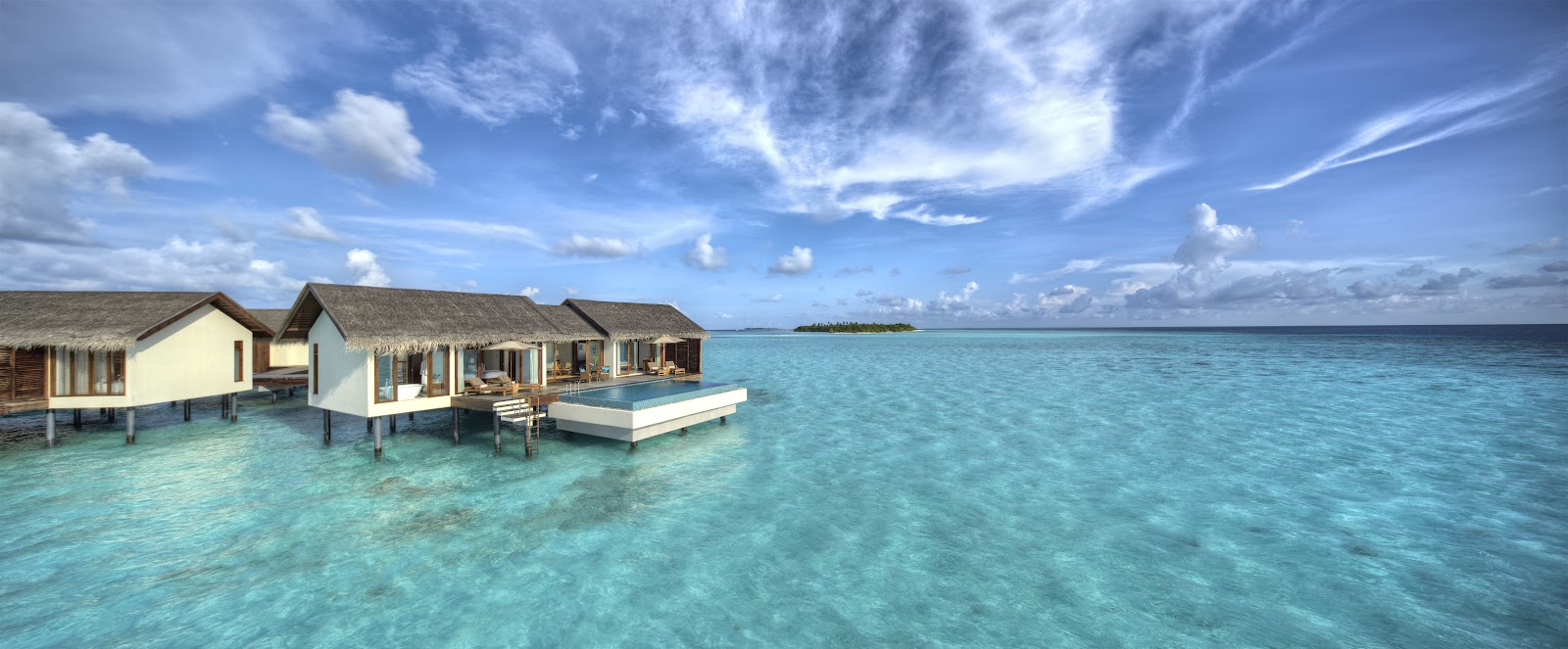 Overwater bungalows with thatched roofs at The Residence Maldives at Falhumaafushi, surrounded by turquoise lagoon water and blue sky.