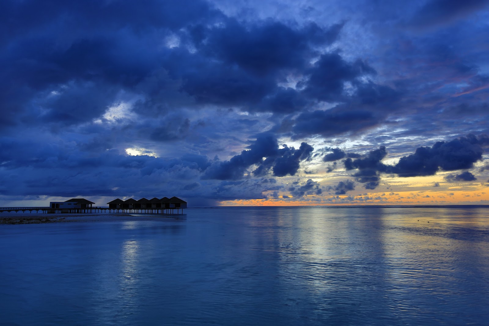 Row of overwater bungalows with thatched roofs at The Residence Maldives, set in clear turquoise lagoon water under blue sky.