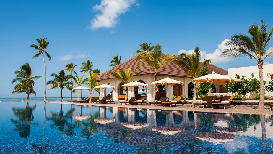 Infinity pool with tiled floor reflects palm trees and a beachfront pavilion with tan umbrellas and lounge chairs under clear blue sky.