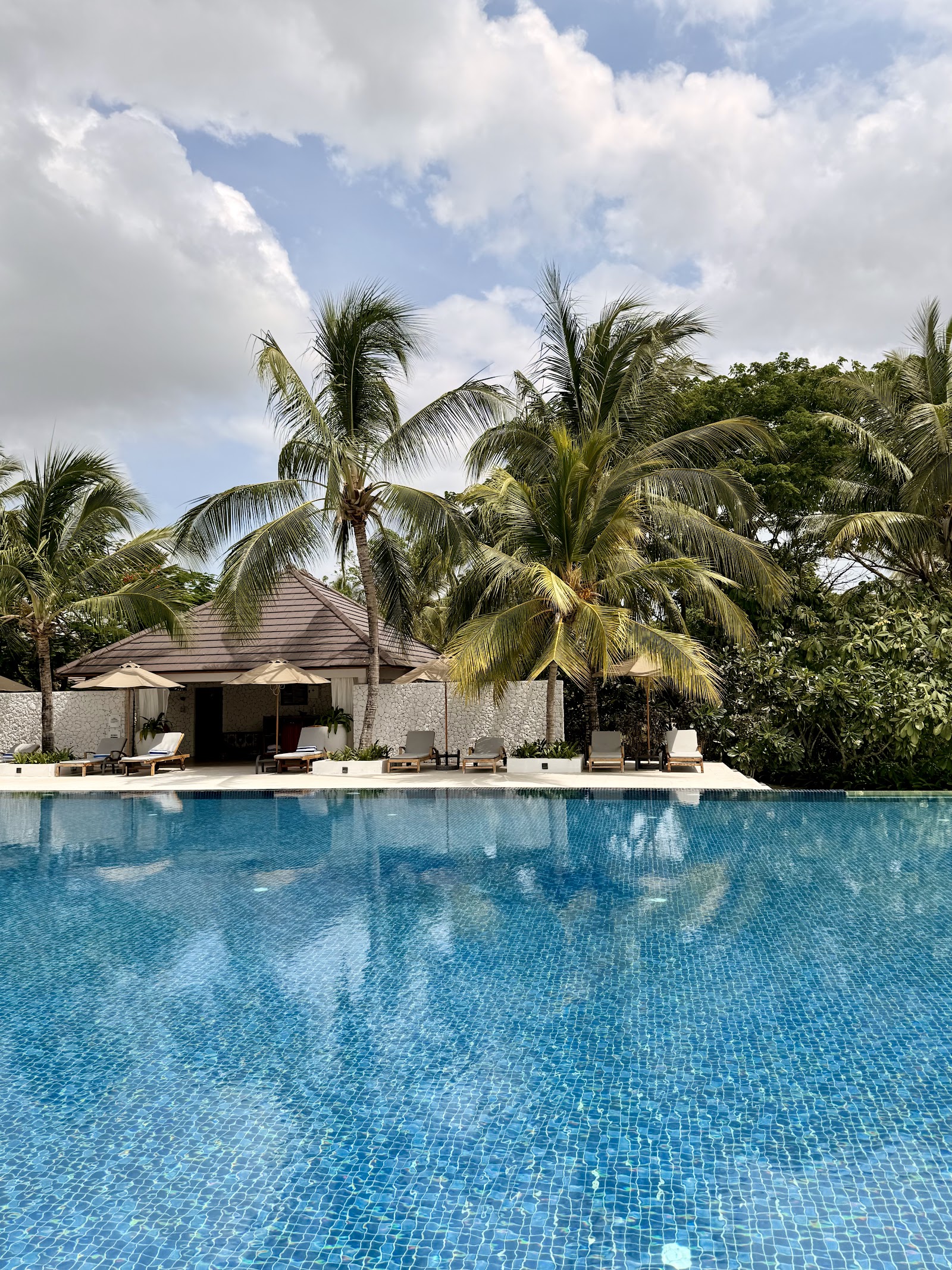 Pool with blue tiles stretches toward a white building with umbrellas and lounge chairs, surrounded by tall palm trees at The Residence Zanzibar.