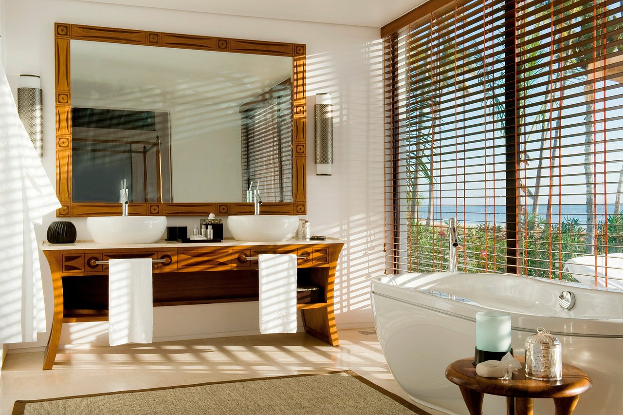 Bathroom with wooden vanity, dual white vessel sinks, large framed mirror, and soaking tub overlooking ocean through slatted wooden shutters at The Residence Zanzibar.