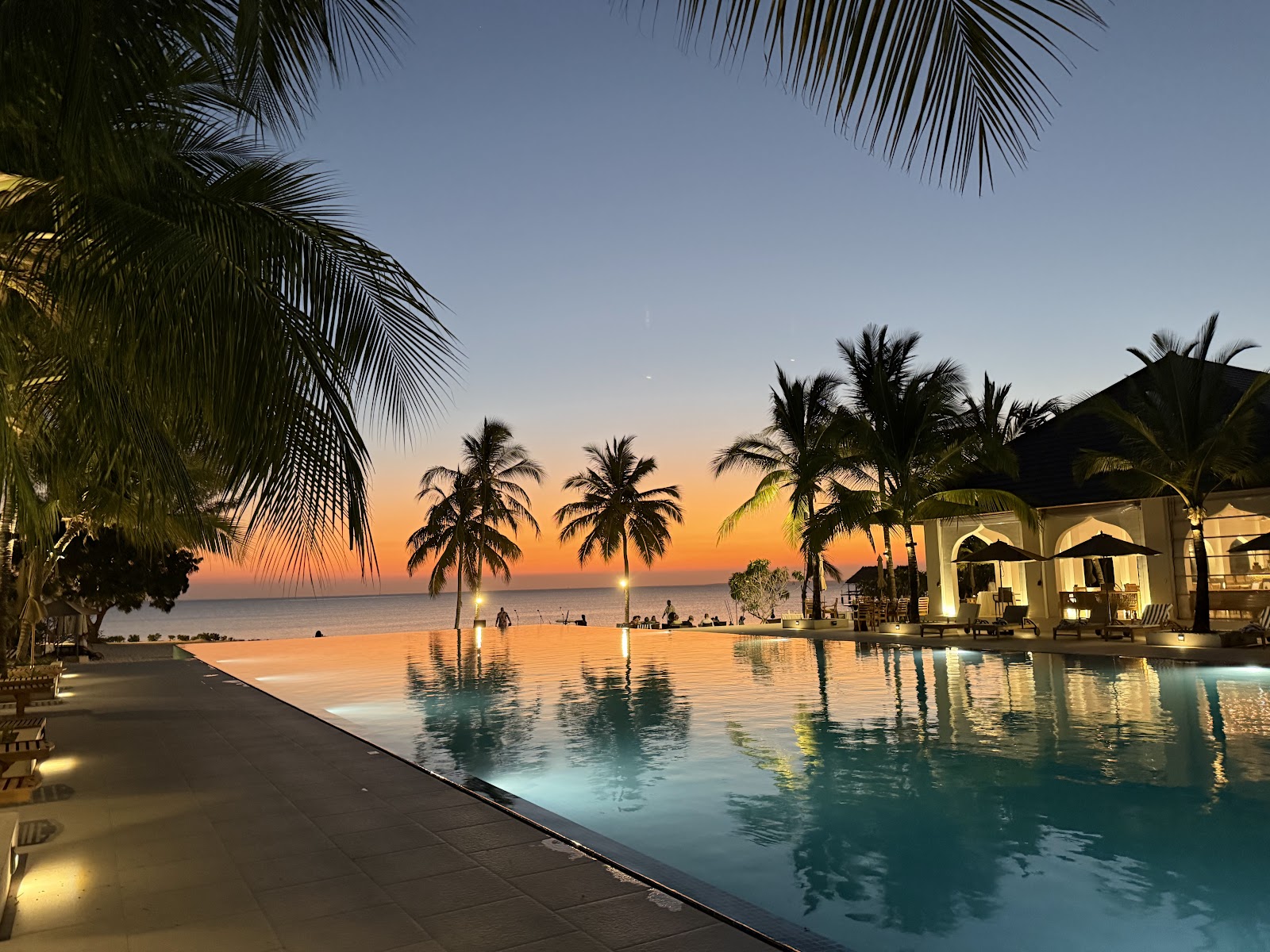 Infinity pool reflecting an orange sunset with palm trees, beach, and illuminated pavilions at The Residence Zanzibar.