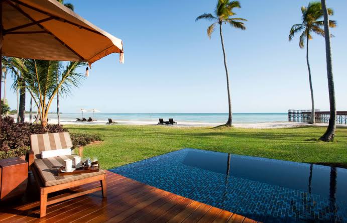 Palm trees and flowering shrubs line a sandy beach with turquoise water and wooden loungers at The Residence Zanzibar.