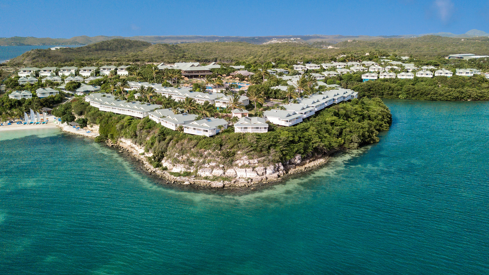 Aerial view of The Verandah Antigua resort nestled on a peninsula surrounded by turquoise water, with white buildings, palm trees, and forested hills beyond.