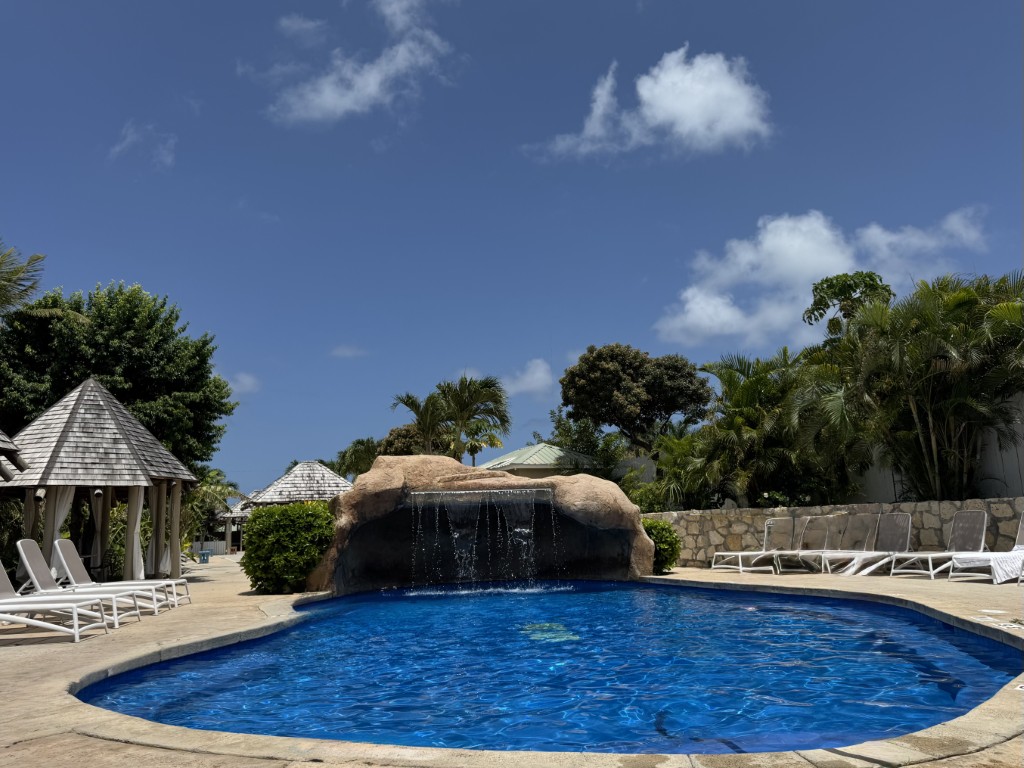 Blue oval pool with rock waterfall feature, white lounge chairs, gazebo, and palm trees under clear sky at The Verandah Antigua.