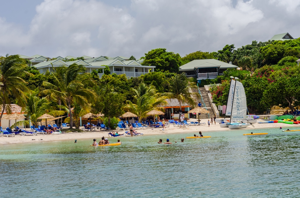 Beach scene at The Verandah Antigua with guests swimming and kayaking in turquoise water, thatched umbrellas on sand, and resort buildings nestled in tropical vegetation.