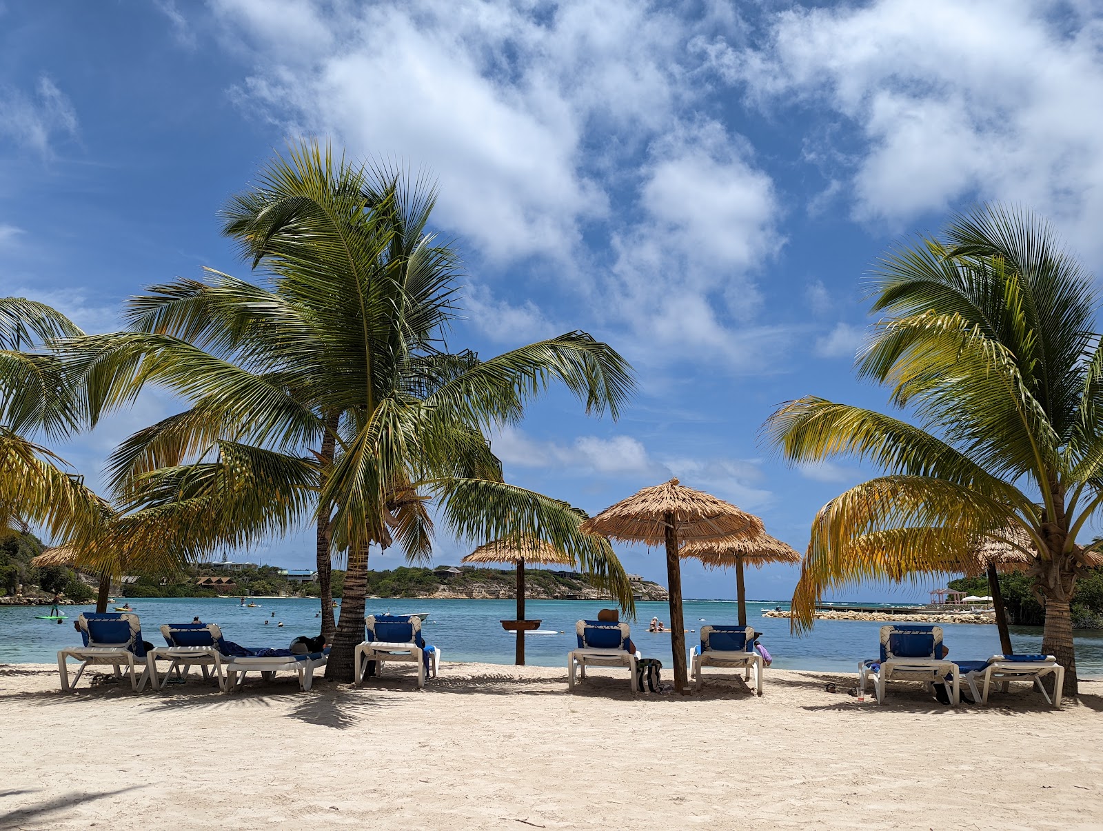 Beach lined with lounge chairs and thatched umbrellas beneath palm trees at The Verandah Antigua, with turquoise water and rocky islands beyond.