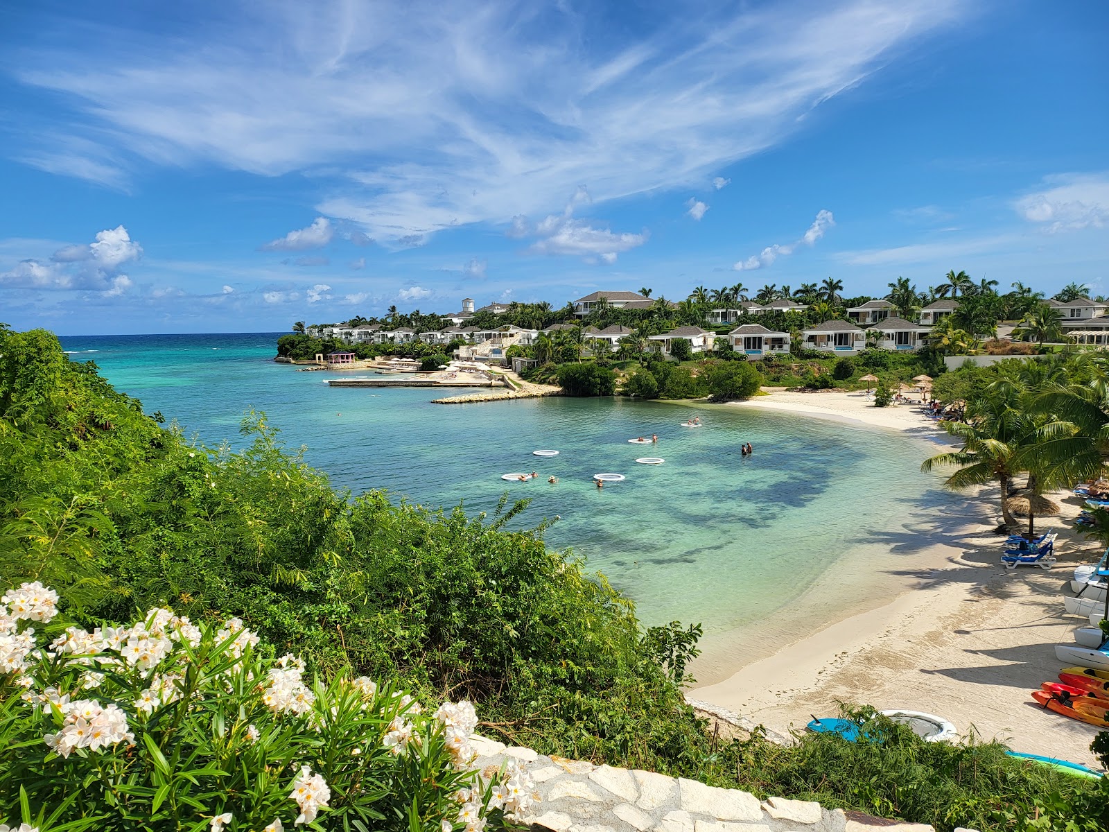 White flowers frame a sandy beach with turquoise water, boats, and swimmers at The Verandah Antigua, with palm-lined villas on the hillside beyond.
