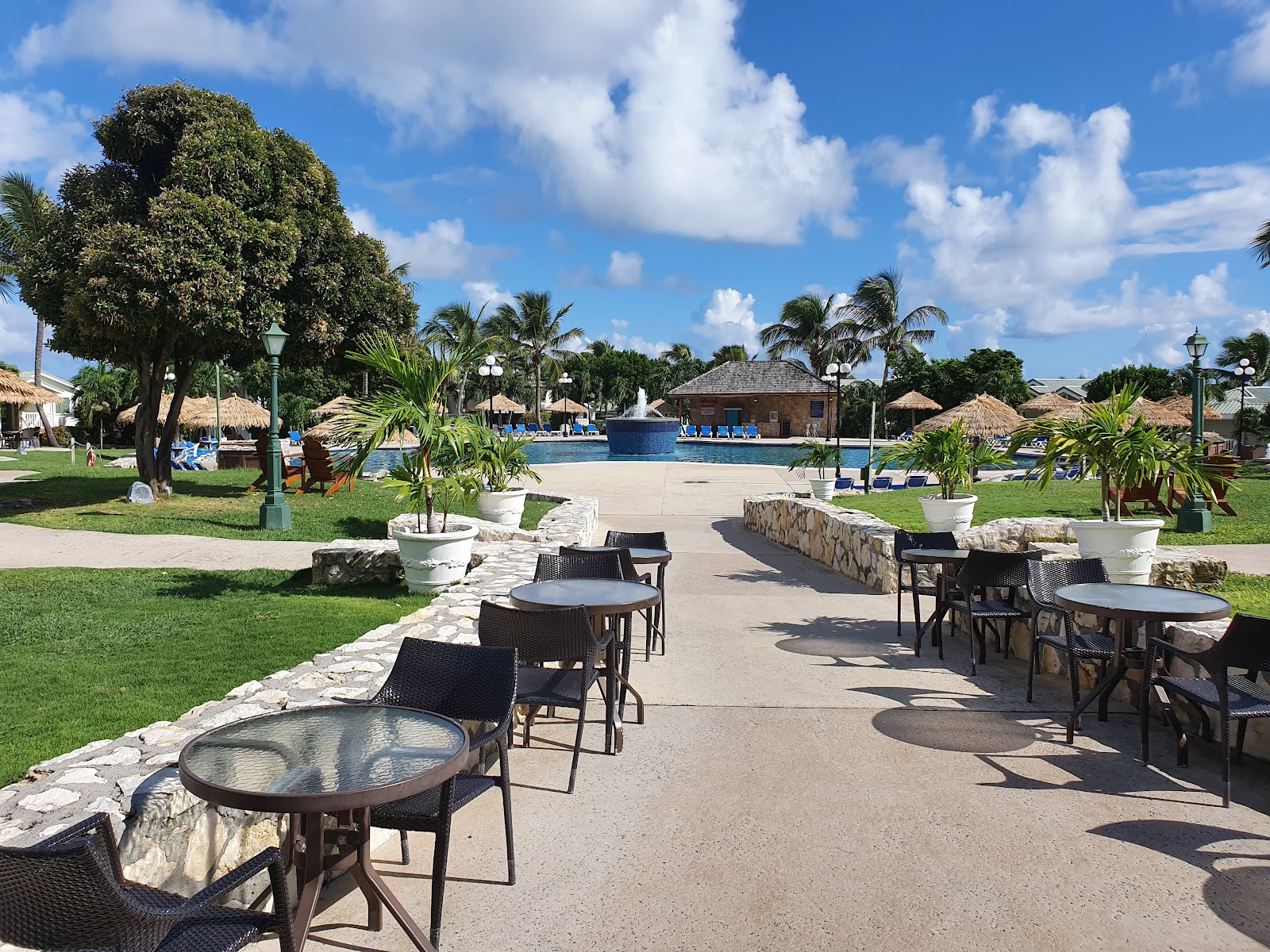 Patio seating area at The Verandah Antigua overlooking a pool, manicured lawn, and thatched-roof structures under blue sky.