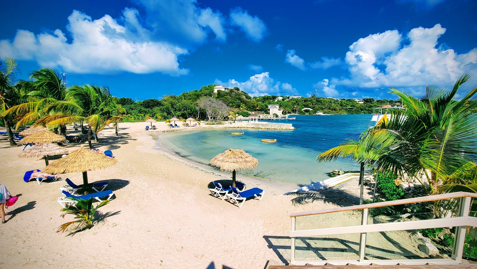 Beach lined with thatched umbrellas and blue loungers overlooking turquoise water, palm trees, and hillside buildings at The Verandah Antigua.