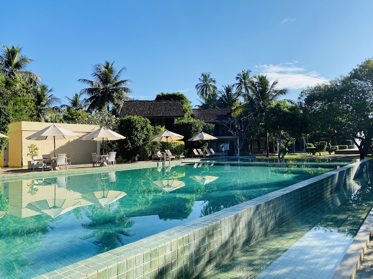 The Villas Wadduwa by Amaya's turquoise pool with tiled sunbathing areas, cream umbrellas, and traditional dark-roofed buildings surrounded by palm trees under clear sky.