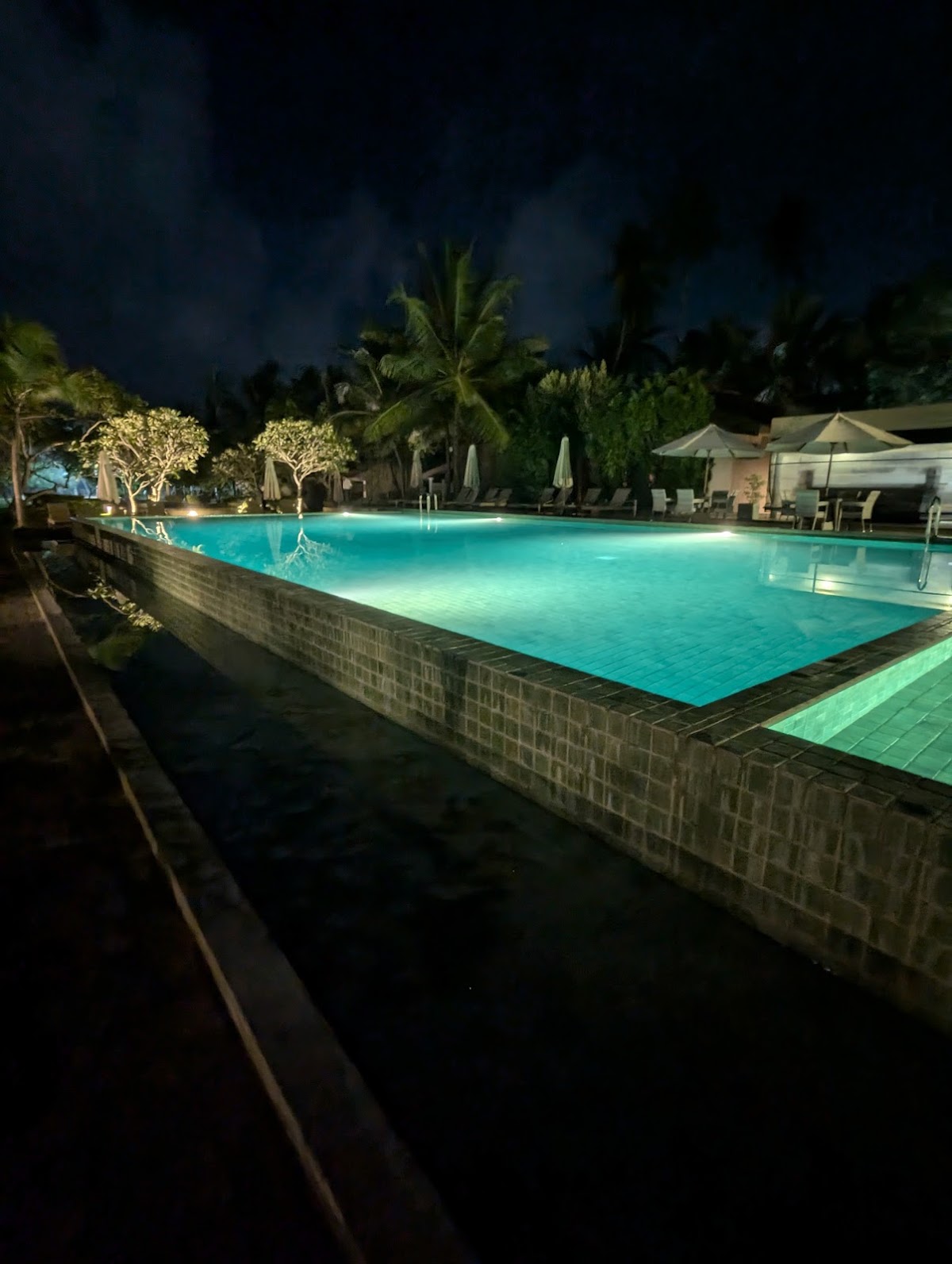 Illuminated resort pool at night surrounded by palm trees, lounge chairs, and umbrellas at The Villas Wadduwa by Amaya.
