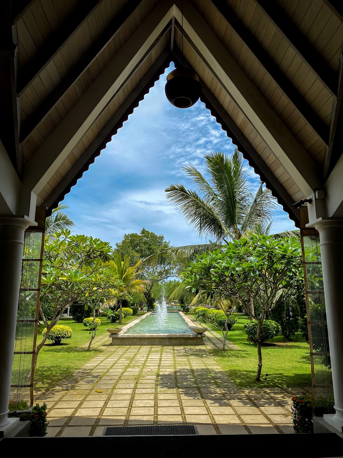View through a peaked wooden doorway of a manicured garden with a central fountain, palm trees, and landscaped grounds at The Villas Wadduwa by Amaya.