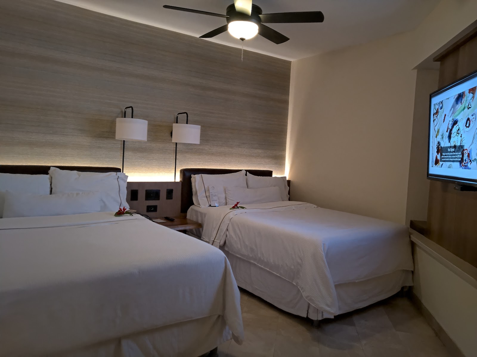 Hotel room with two queen beds, white linens, wall-mounted lamps, ceiling fan, and flat-screen TV.