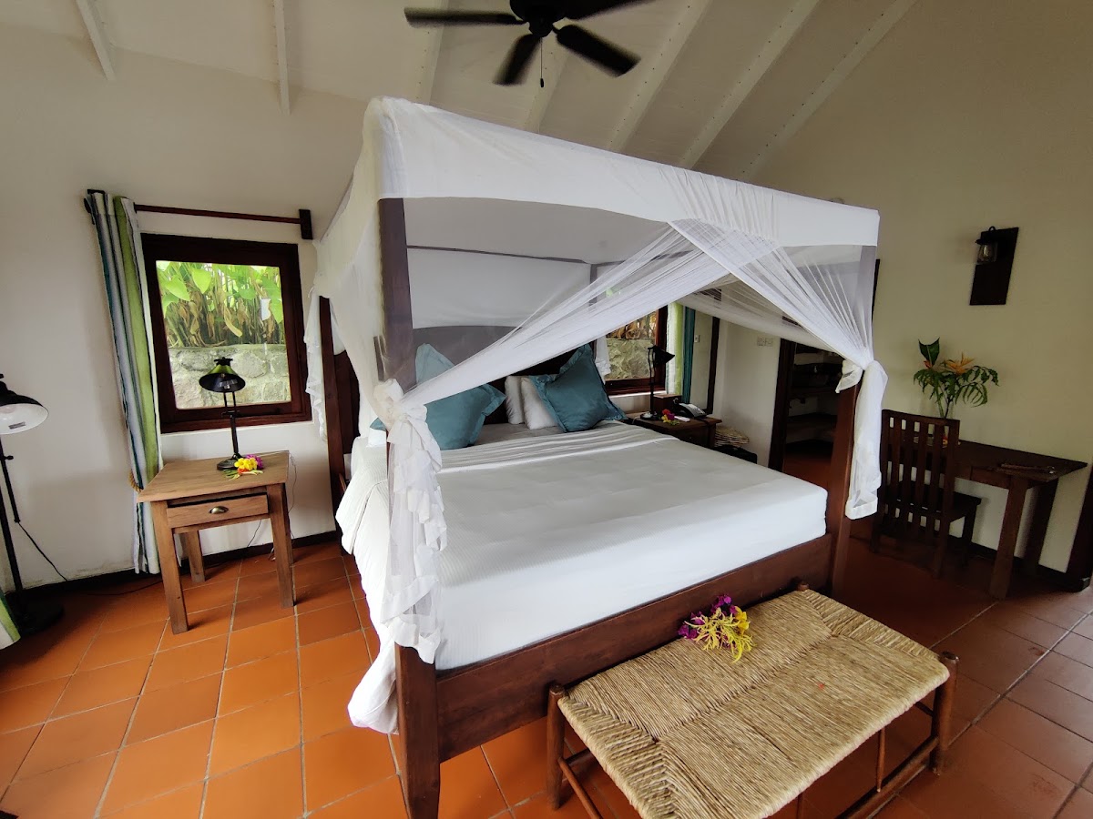 Four-poster canopy bed with white drapes and teal pillows in a guest room with terracotta tile floor and tropical garden view.