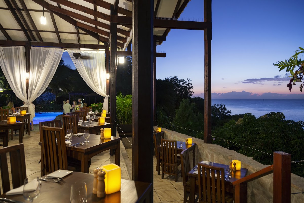Ti Kaye Resort & Spa dining terrace at dusk with wooden tables, white curtains, and ocean view beyond tropical vegetation.