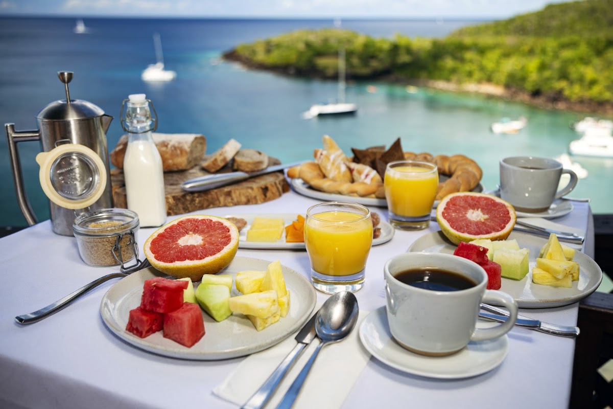 Breakfast table with fresh fruit, pastries, and coffee overlooking turquoise water and moored boats at Ti Kaye Resort & Spa.