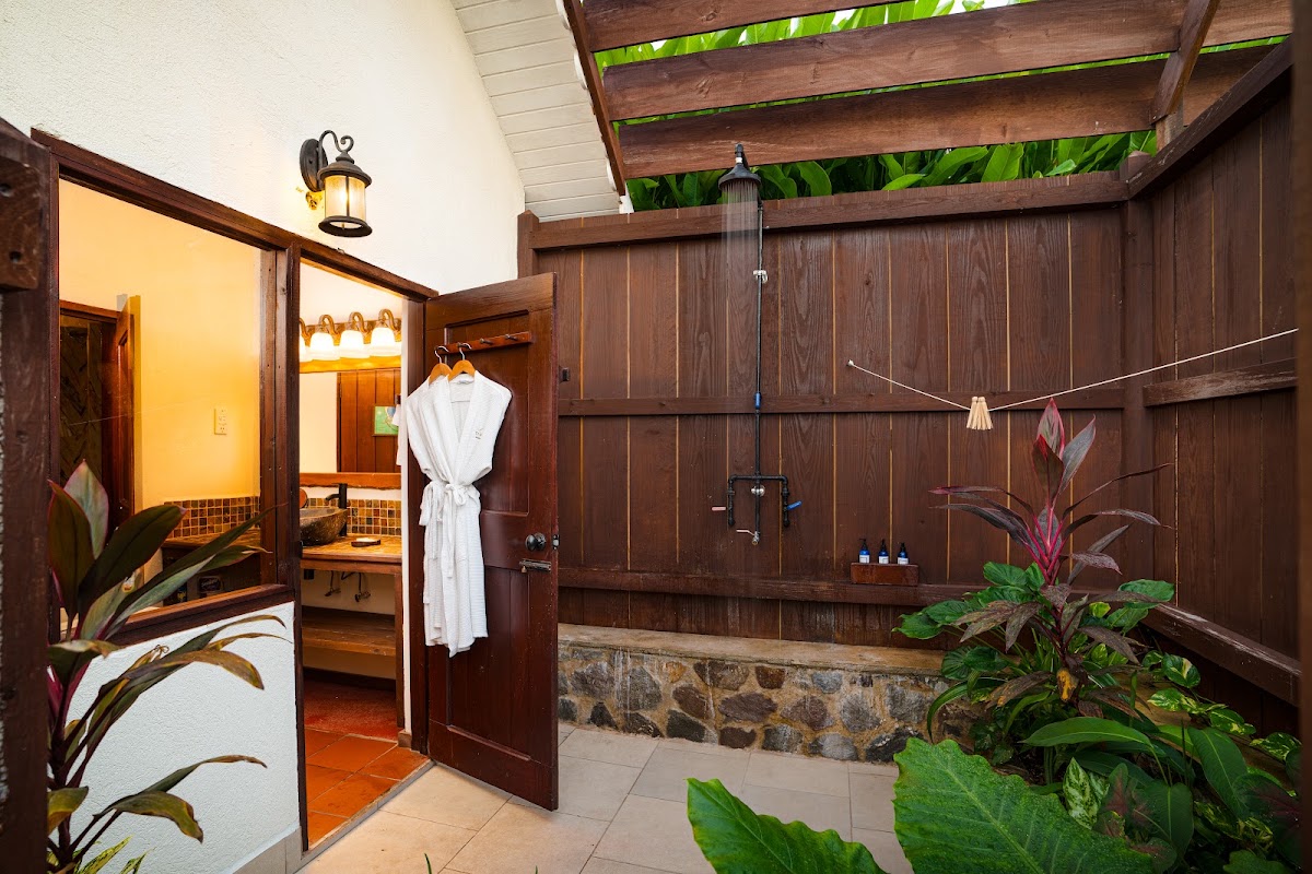Open-air outdoor shower with dark wood paneling, stone base, hanging white robe, tropical plants, and overhead lattice pergola at Ti Kaye Resort & Spa.