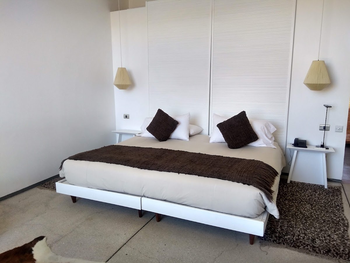 Modern bedroom at Tierra Atacama with king bed, dark throw pillows, white bedside tables, and pendant lamps flanking a textured headboard.