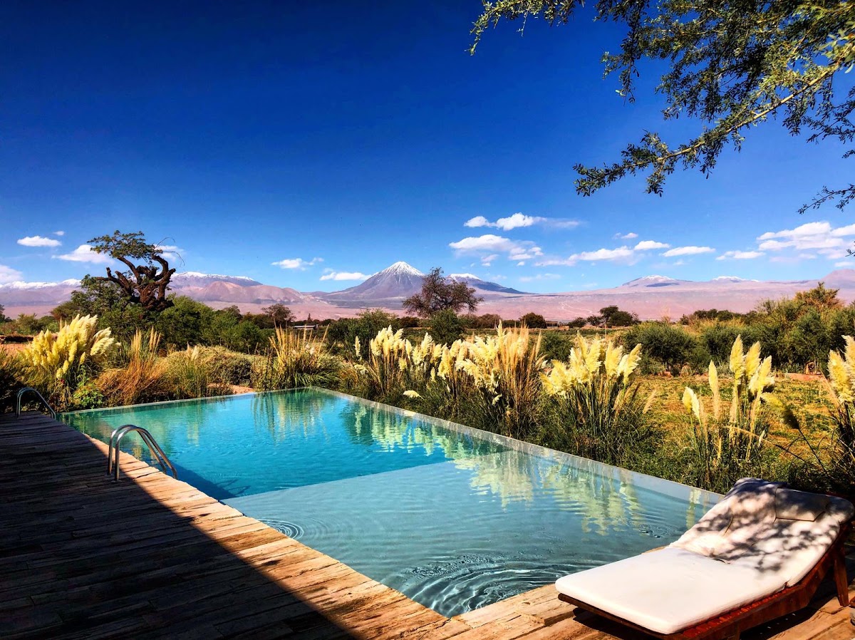 Resort pool with wooden deck overlooks desert landscape, native grasses, and snow-capped volcano in the distance under clear blue sky.