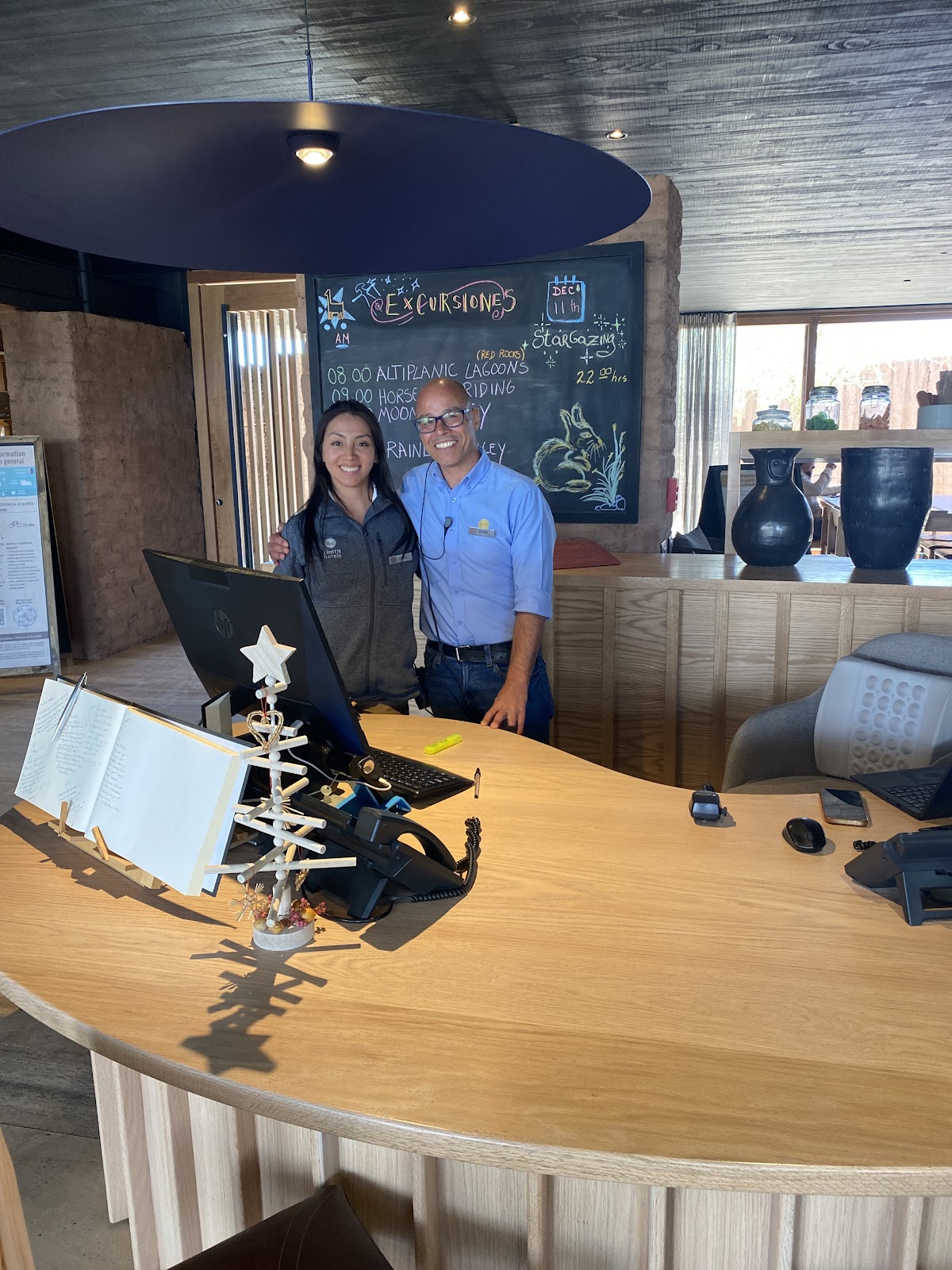 Two staff members stand at a wooden reception desk with a computer and activity board listing excursions behind them.