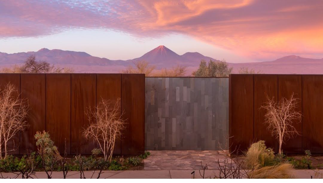 Rusted metal gates and sparse desert vegetation frame a snow-capped volcano at sunset under a pink and orange sky at Tierra Atacama.