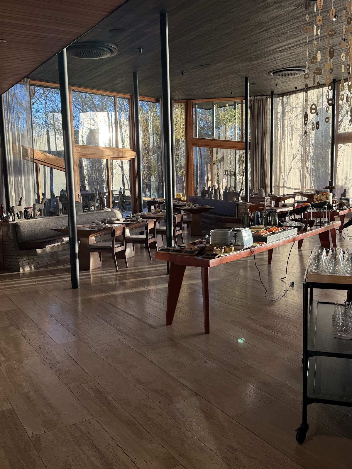 Modern dining room with wooden floors and large windows overlooking bare trees, featuring dark wooden tables and hanging brass ring decorations.