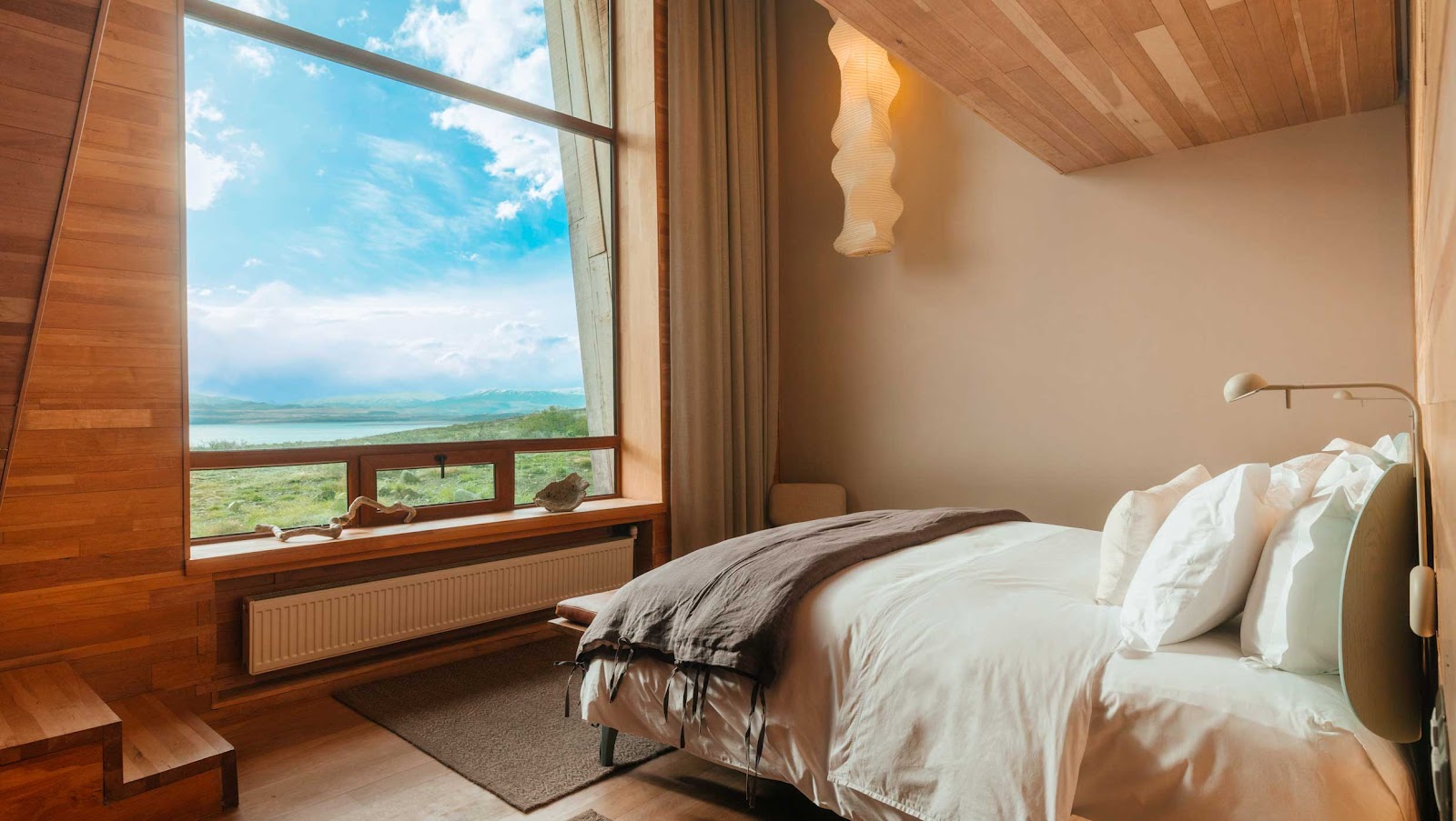 Bedroom at Tierra Patagonia with wooden interior, large window overlooking Patagonian landscape and lake under blue sky.